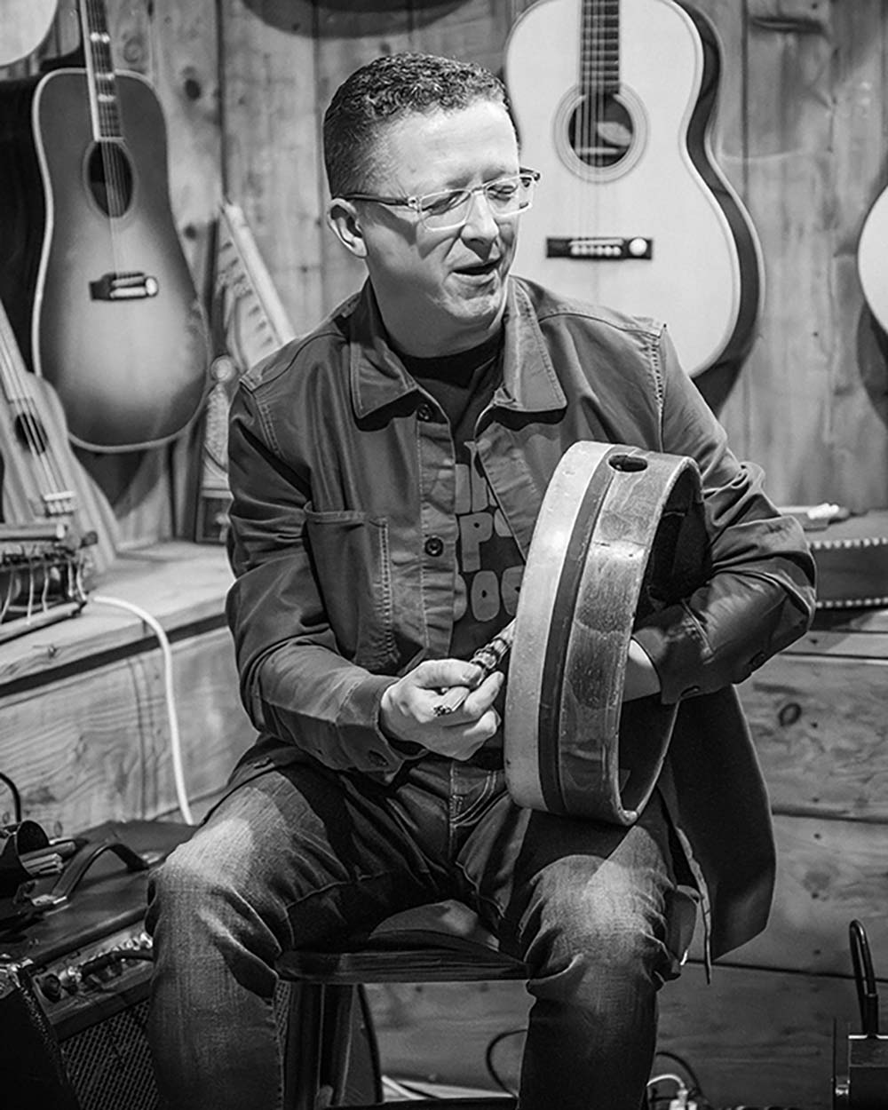 Black and white picture of Brian Morrissey playing the bodhran. He is in a seated postion. There are musical instruments hanging up in the background