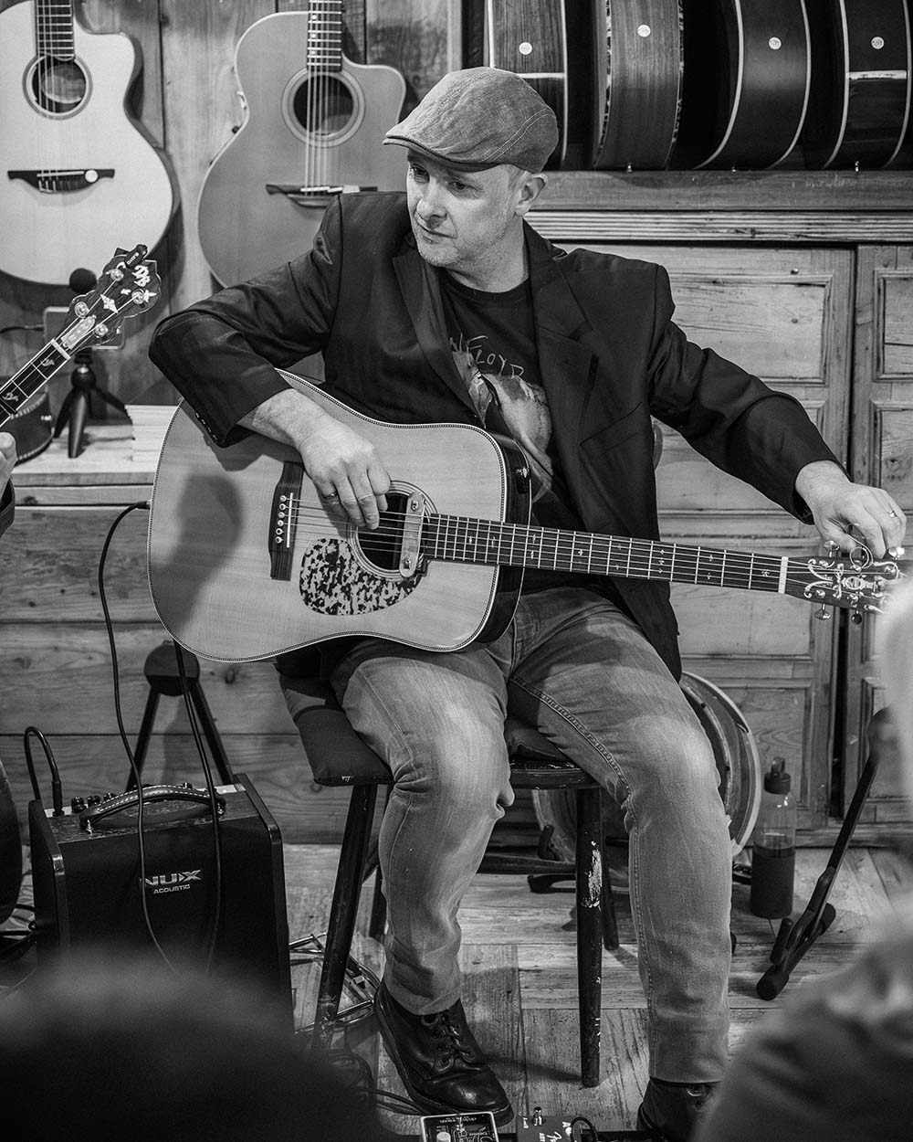 Black and white picture of Eoin Coughlan playing the guitar. He is in a seated position on a stage with musical instruments hanging behind him.
