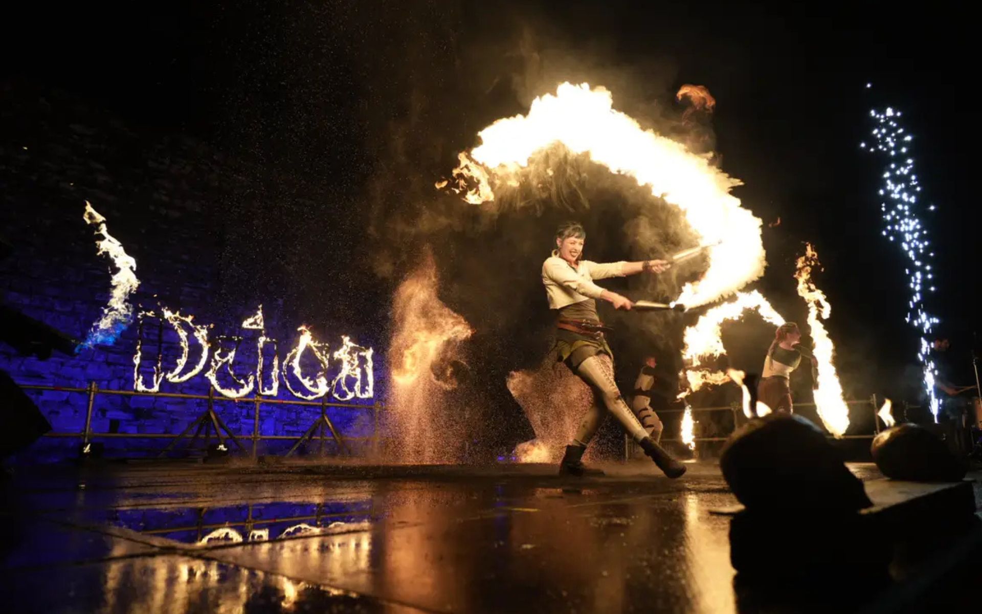 A flame thrower performs in the foreground with the word PUCA in flames behind her.